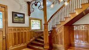 Eine imposante Treppe mit Holllaufstegen und einem Chandelier, die zu einer Empore mit einem gerahmten Landschaftsbild und einem Buntglasfenster führt.