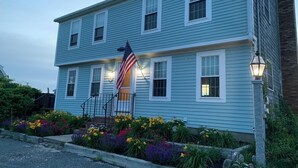 A two-story building with a flag in front, a front porch, and a flower bed.