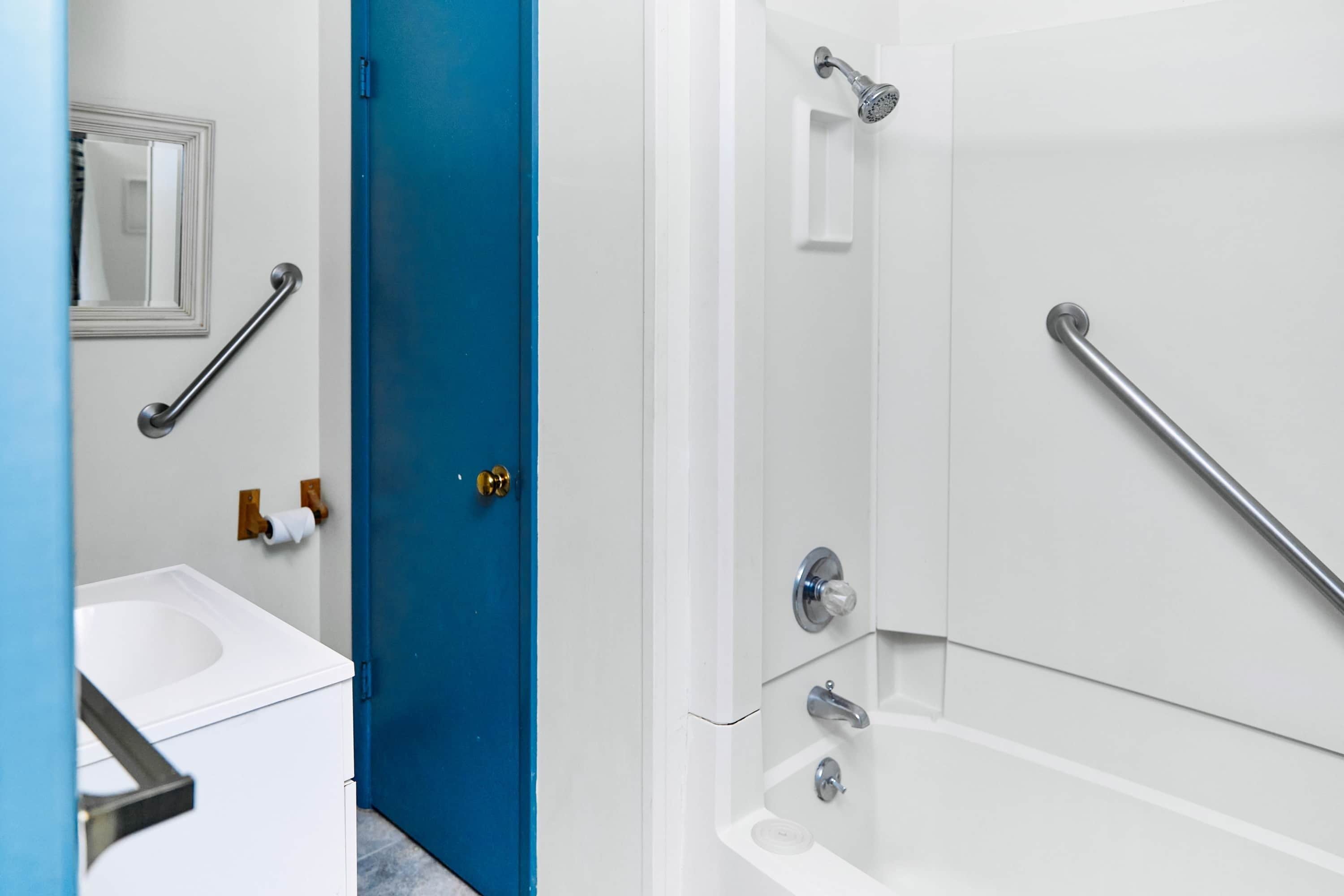 Une salle de bain avec un lavabo blanc, une porte bleue et une douche équipée de barres d’appui.