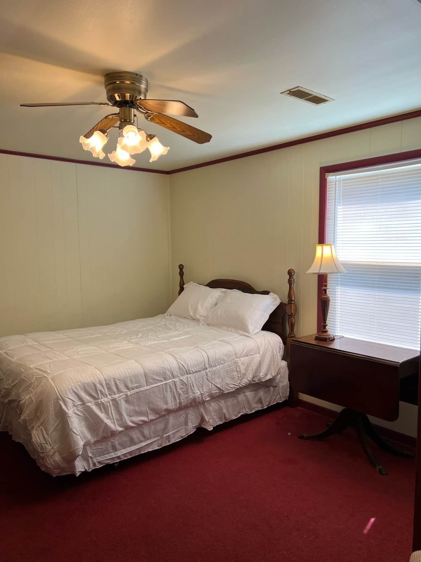 A bedroom with a bed, a desk, a lamp, a ceiling fan, and a window with blinds.