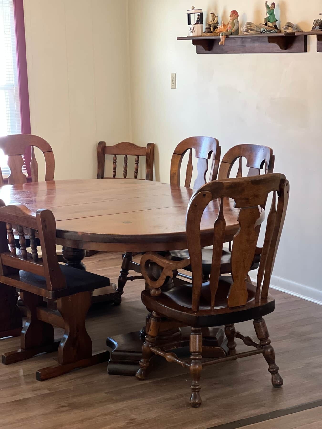 A dining room with a wooden table and chairs, a shelf with figurines, and a window with blinds.