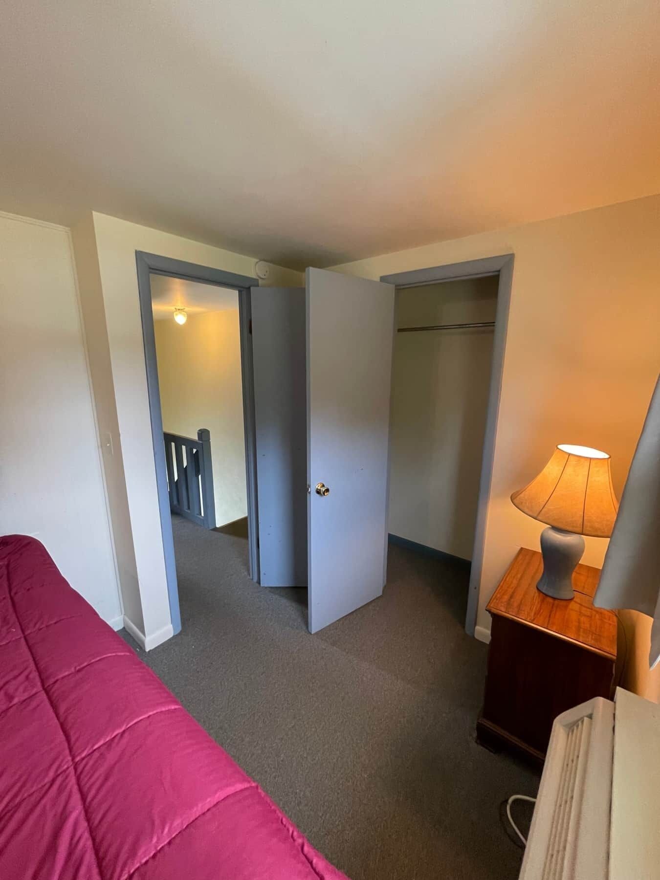 A hotel room with a bed, a nightstand with a lamp, a closet, and a door leading to another room.
