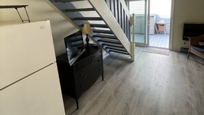 A spacious living area with a wooden floor, a black sofa, a lamp, and a TV. A staircase leads to an upper level.