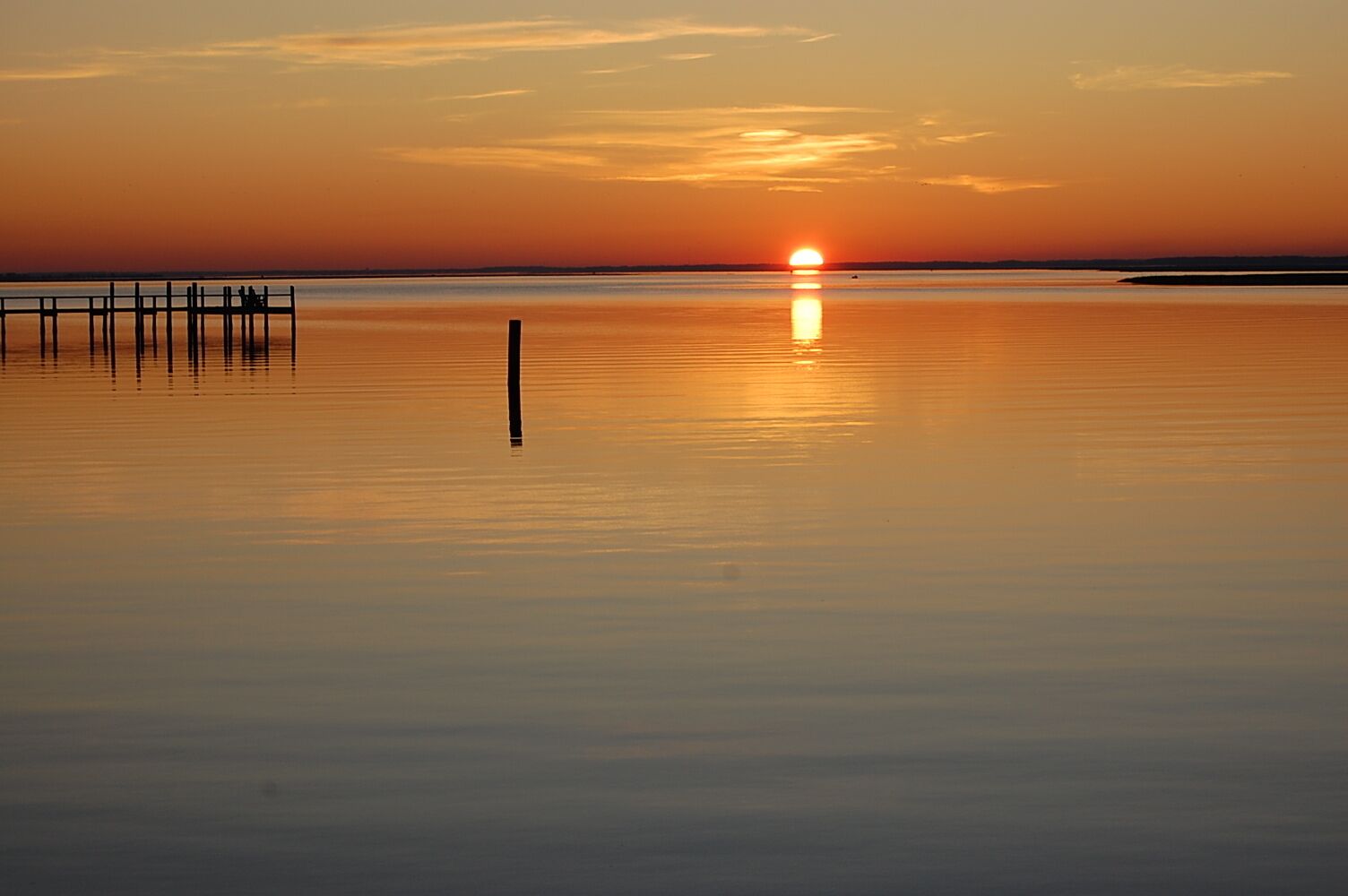 Une étendue d’eau calme, avec une jetée en bois qui s’avance sur l’eau et dans laquelle se reflète le coucher de soleil.