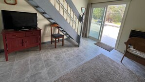 A spacious living room with a large staircase, a television on a cabinet, and a view of the outdoors through a glass door.