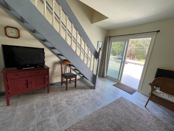 A spacious living room with a large staircase, a television on a cabinet, and a view of the outdoors through a glass door.