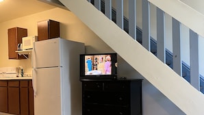 Una cocina con refrigerador, microondas y armarios de madera. En primer plano, un mueble bajo para televisión negro con televisor de pantalla plana.