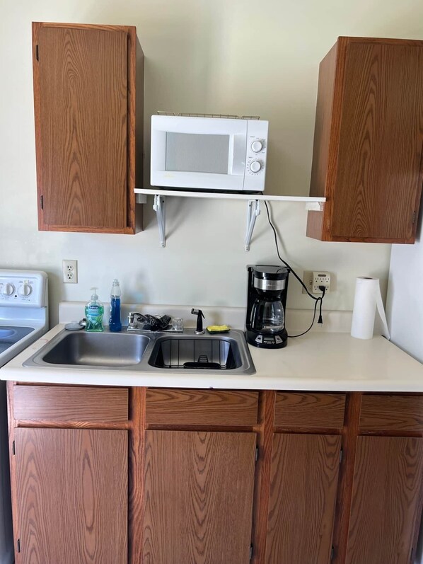 A compact kitchen with wooden cabinets, a white microwave, a coffee maker, a sink, and a stove. - Sunset Villa (Chincoteague)