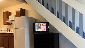 A kitchen with a refrigerator, microwave, and wooden cabinets. A black TV stand with a flat-screen TV is in the foreground.