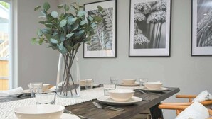 Een eetruimte met een lange tafel gedekt voor vier personen, zwart-wit foto’s aan de muur en een vaas met groene takken op de tafel.