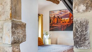 Ein Schlafzimmer mit Steinwand, einem Bett mit gemustertem Überwurf, einem weißen Heizkörper, einer Vase mit Blumen und einem Stadtbild an der Wand.
