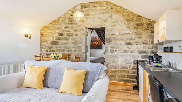 Ein gemütliches Zimmer mit Steinwand, einer Couch mit gelben Kissen, Holzboden und einer Küchenzeile mit Spüle und Schränken.