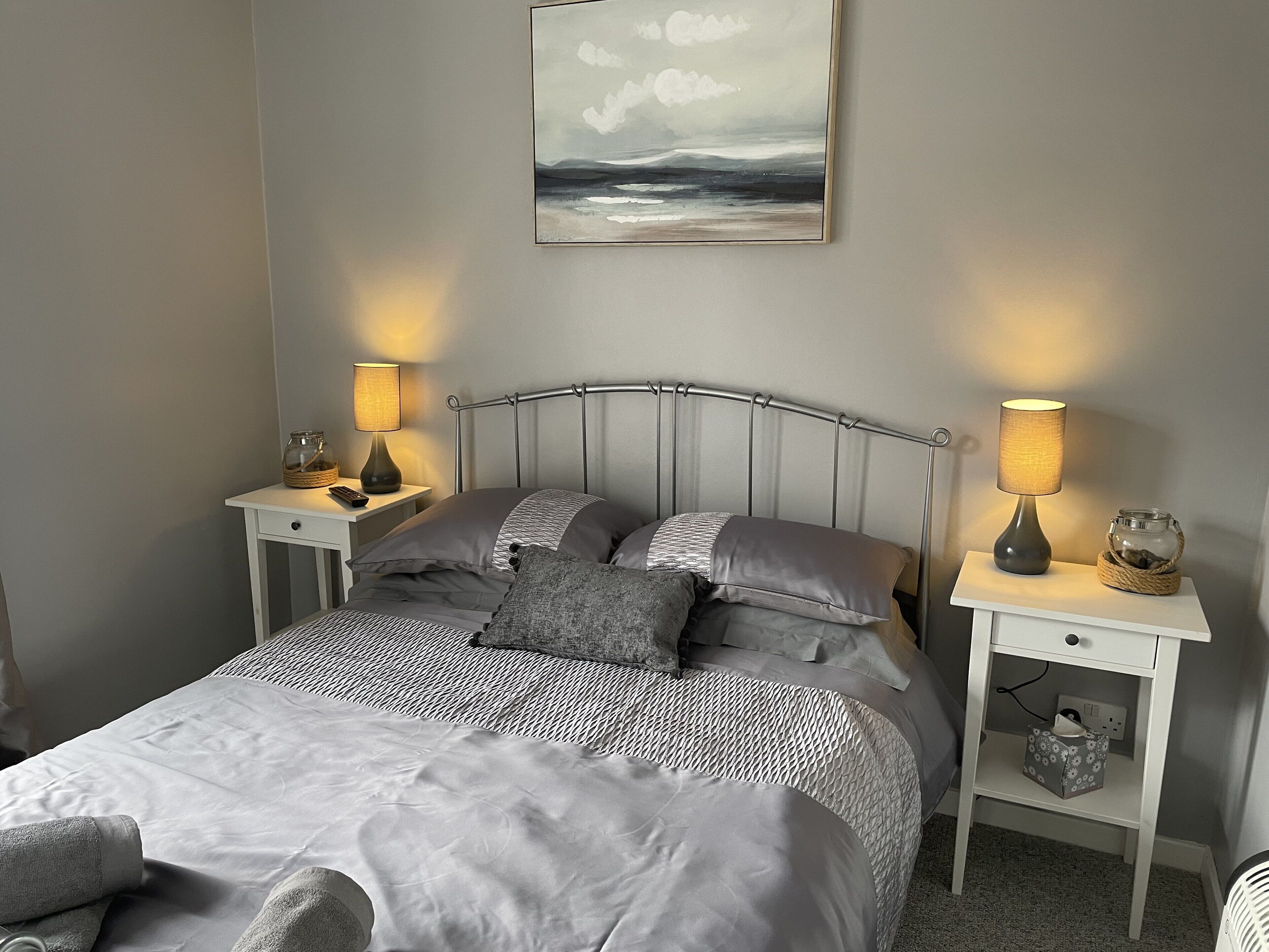 A neatly made bed with grey bedding, two bedside tables with lamps, a wall-mounted painting, and a small bedside table with a jar and a basket.