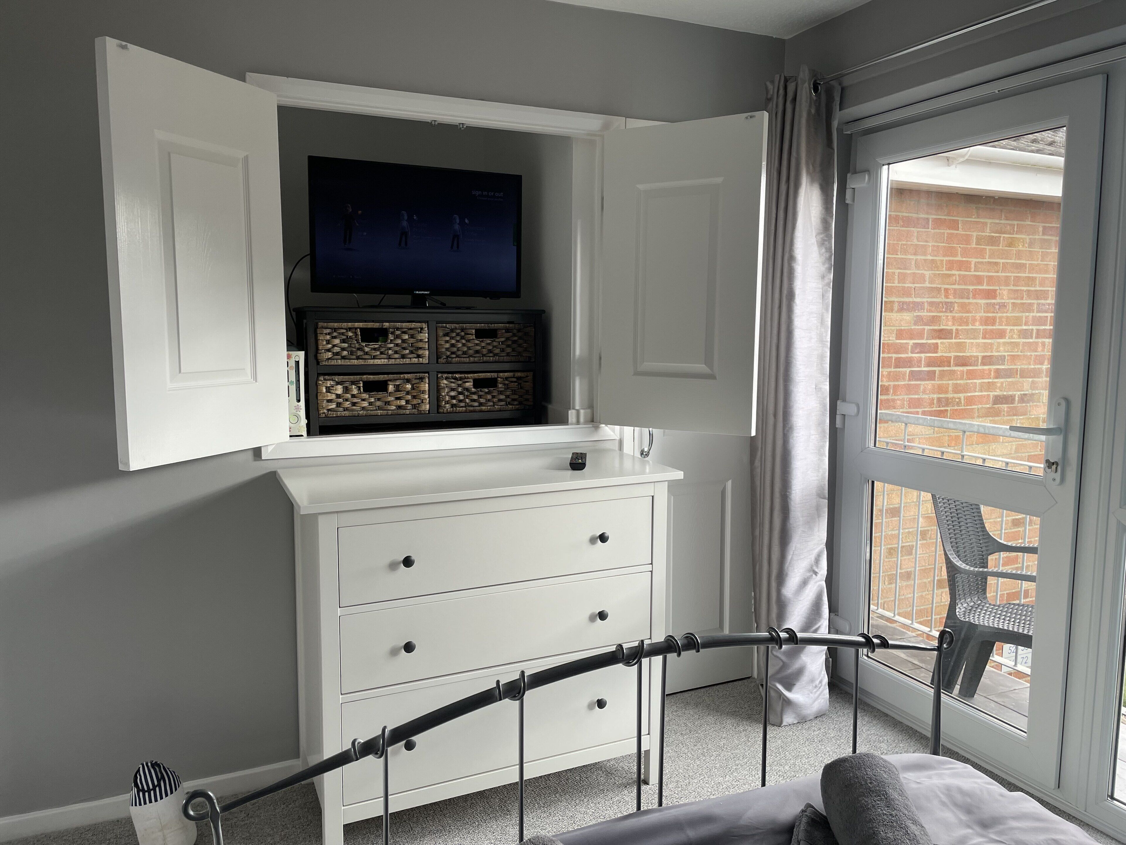 Une chambre avec un lit, une commode blanche, un téléviseur placé au-dessus de la commode, et une fenêtre donnant sur un mur de briques et une chaise à l’extérieur.