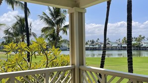 A covered outdoor area with a wooden table and chairs, overlooking a grassy area and a body of water with palm trees.