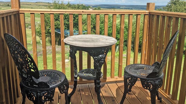Una terraza de madera con una mesa redonda y dos sillas decoradas, con vistas a un paisaje cubierto de hierba.