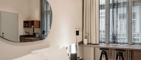 A hotel room with a large bed, a desk with a chair, a mirror, a window with curtains, and a radiator.