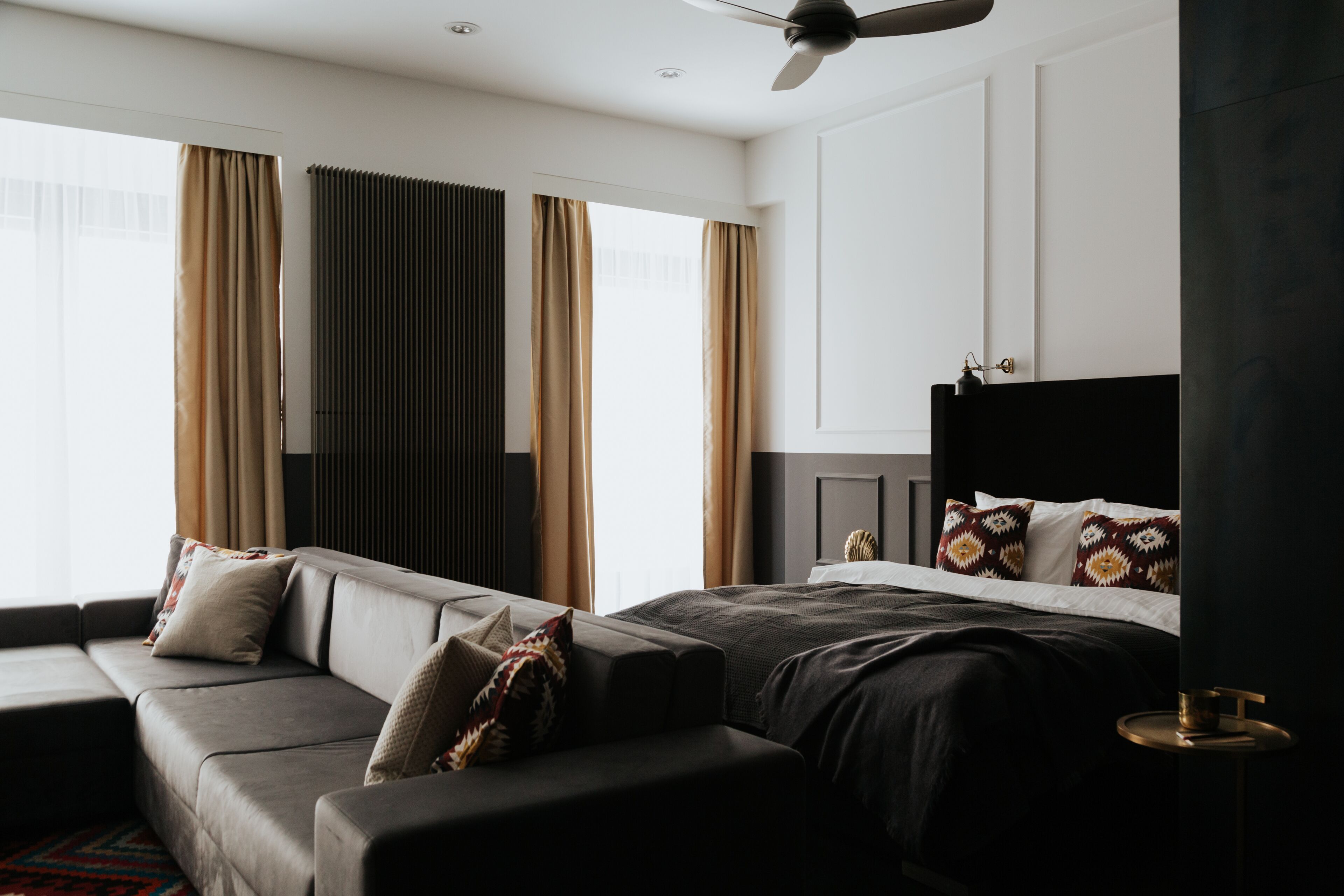 A modern hotel room with a sofa, a bed with patterned pillows, a ceiling fan, and large windows with curtains.