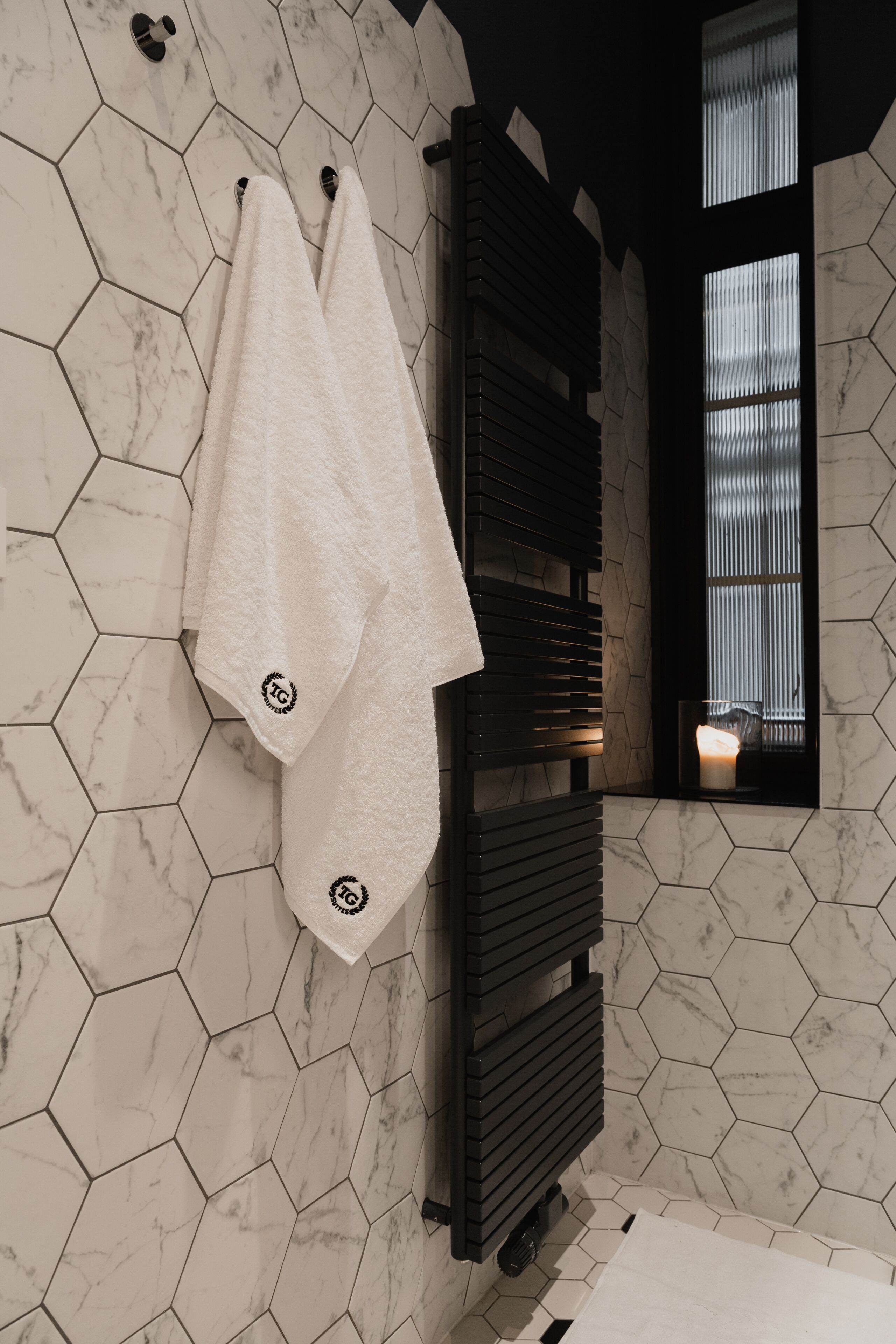 A bathroom with hexagonal marble tiles, a towel rack with two towels, a black towel warmer, and a window with blinds.