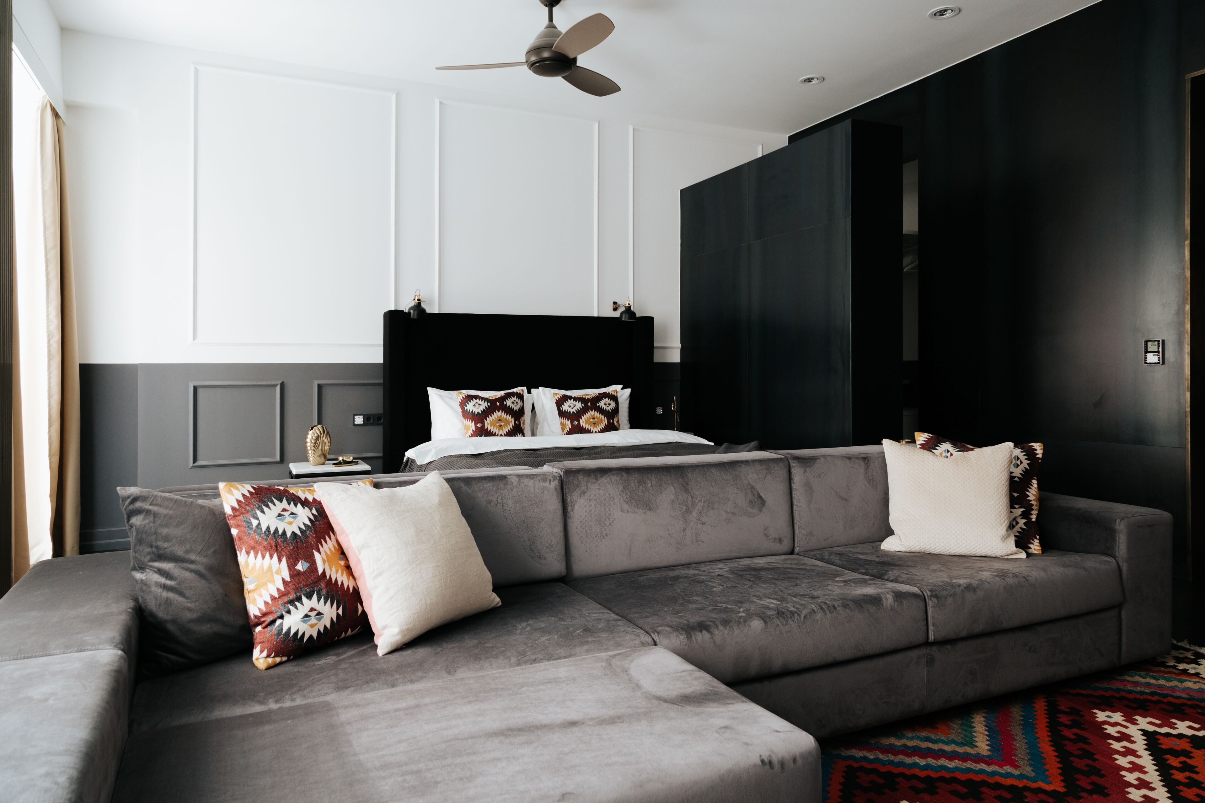 A modern bedroom with a grey sectional sofa, a black headboard bed, and a patterned rug.