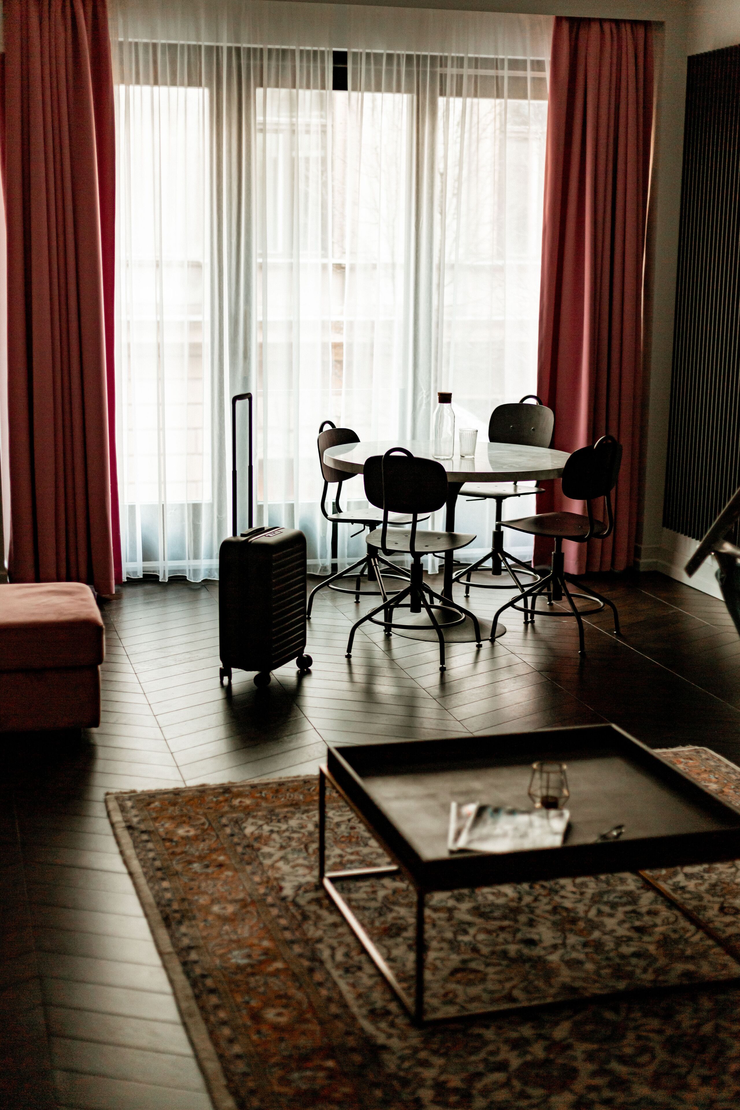 A hotel room with a dining table, four chairs, a small sofa, a coffee table, a suitcase, and a patterned rug.