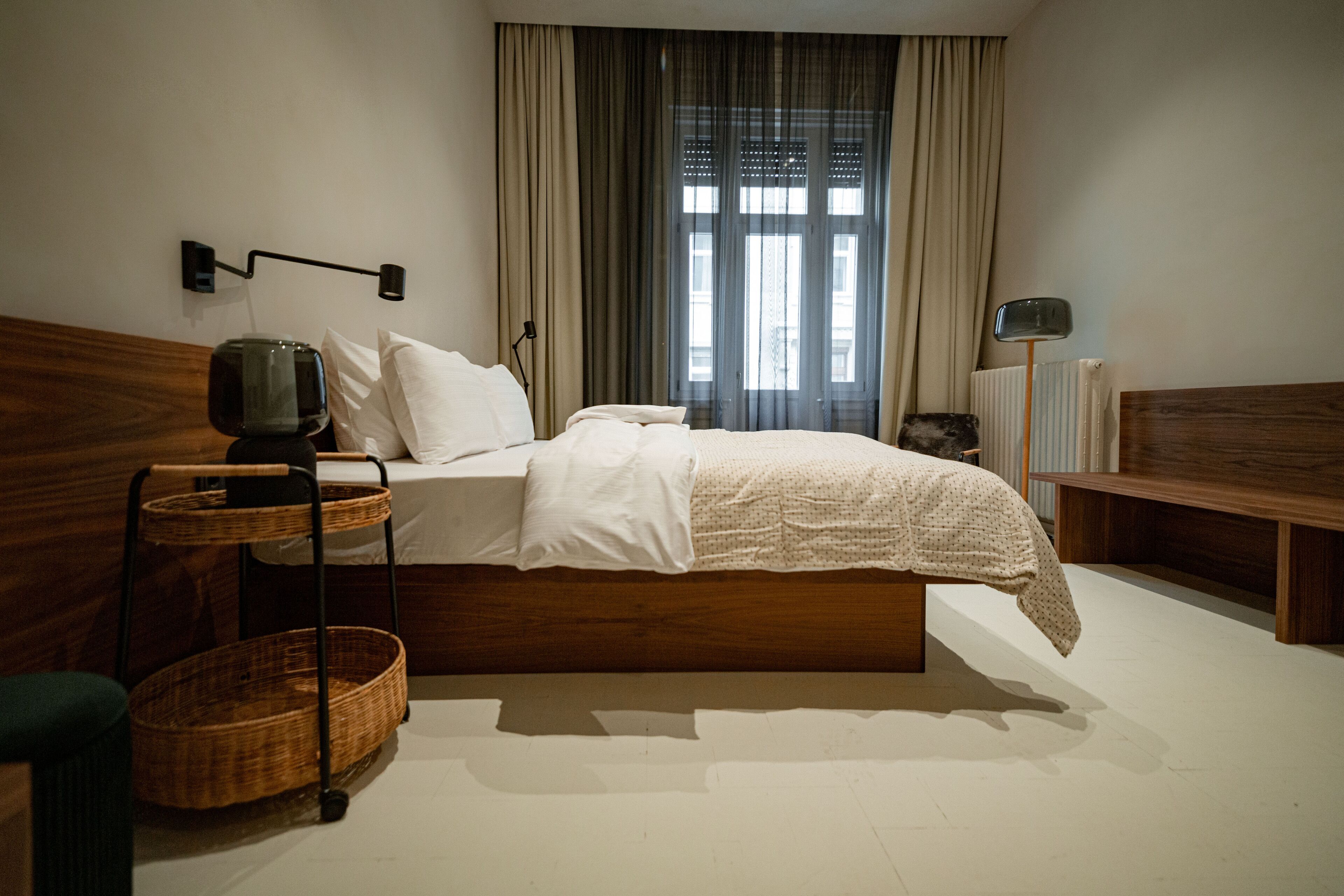 A neatly arranged bedroom with a wooden headboard, a bedside table with a lamp, a bedside basket, and a large window with curtains.
