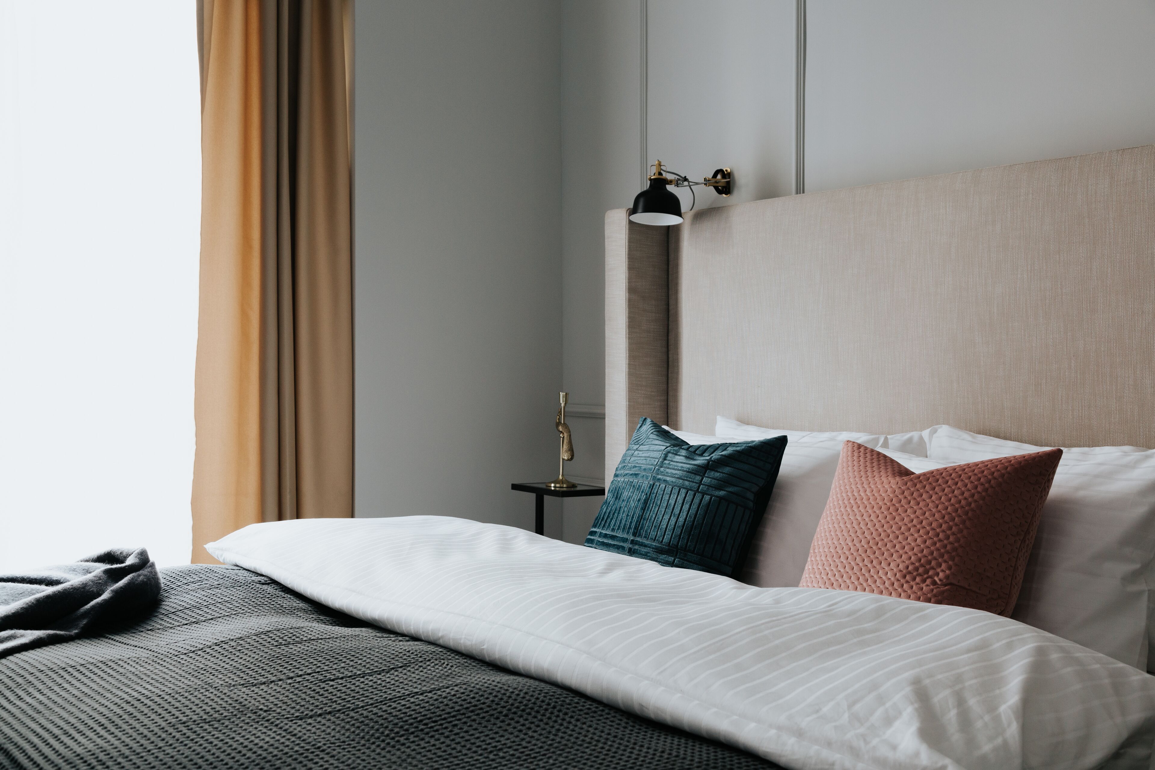 Un lit bien fait, avec une couette blanche, un plaid gris et deux oreillers aux couleurs turquoise et rouille. Une lampe de chevet avec un abat-jour noir est posée sur une petite table à côté du lit.