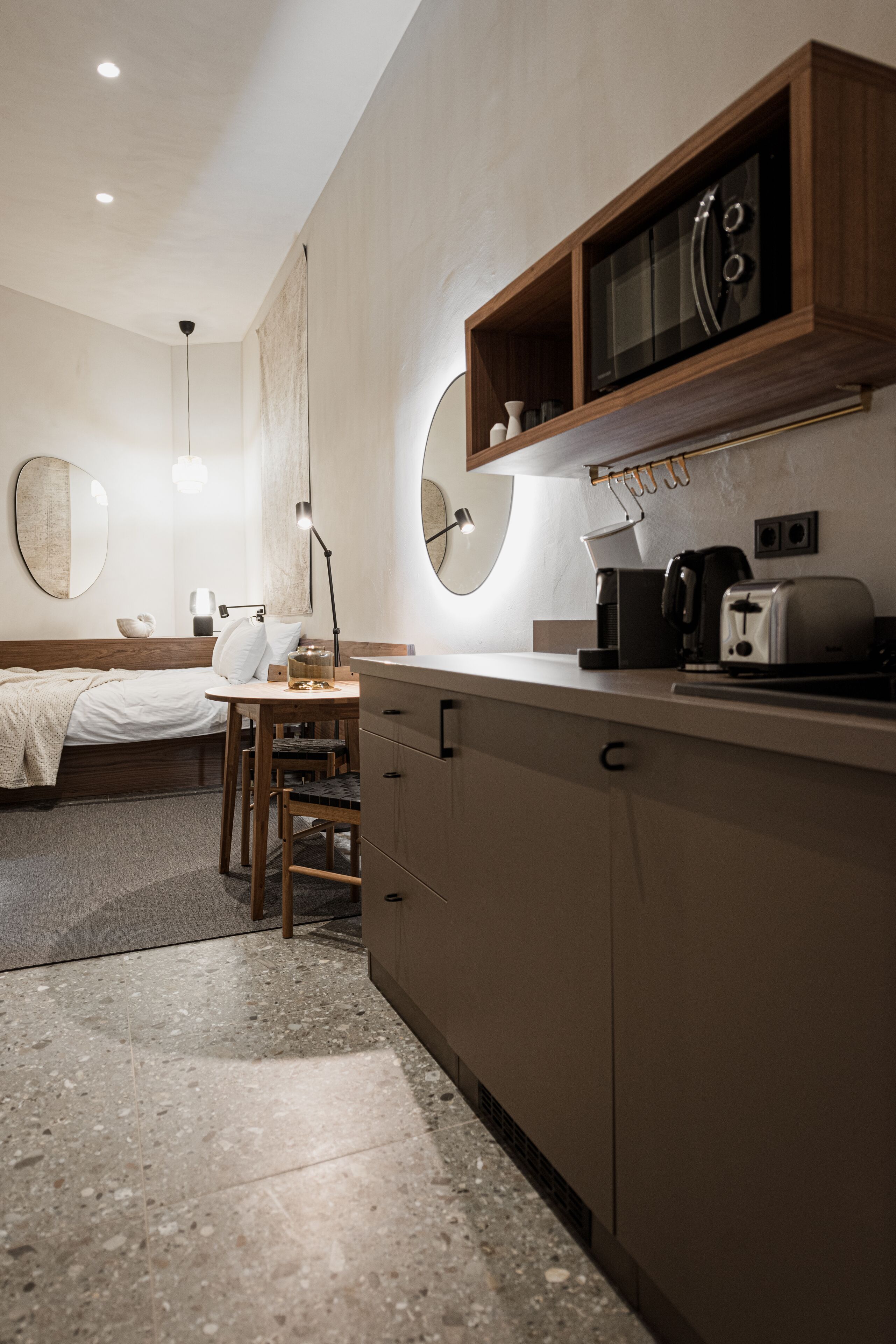 A modern, compact bedroom with a bed, bedside table, and a mirror. Adjacent is a kitchenette with a stove, toaster, and coffee maker.
