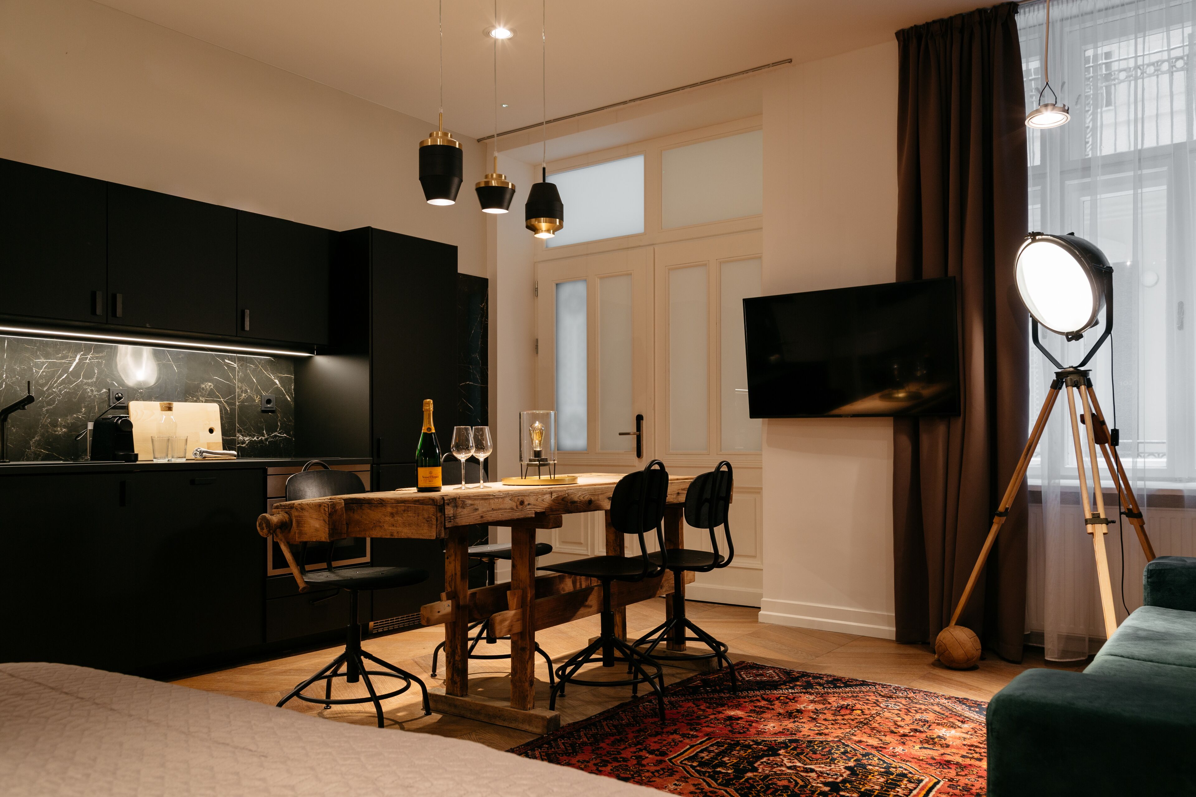 A modern kitchen with dark cabinets, a wooden dining table, and black chairs. There is a tripod with a ring light, a flat-screen TV on the wall, and a large window with curtains.