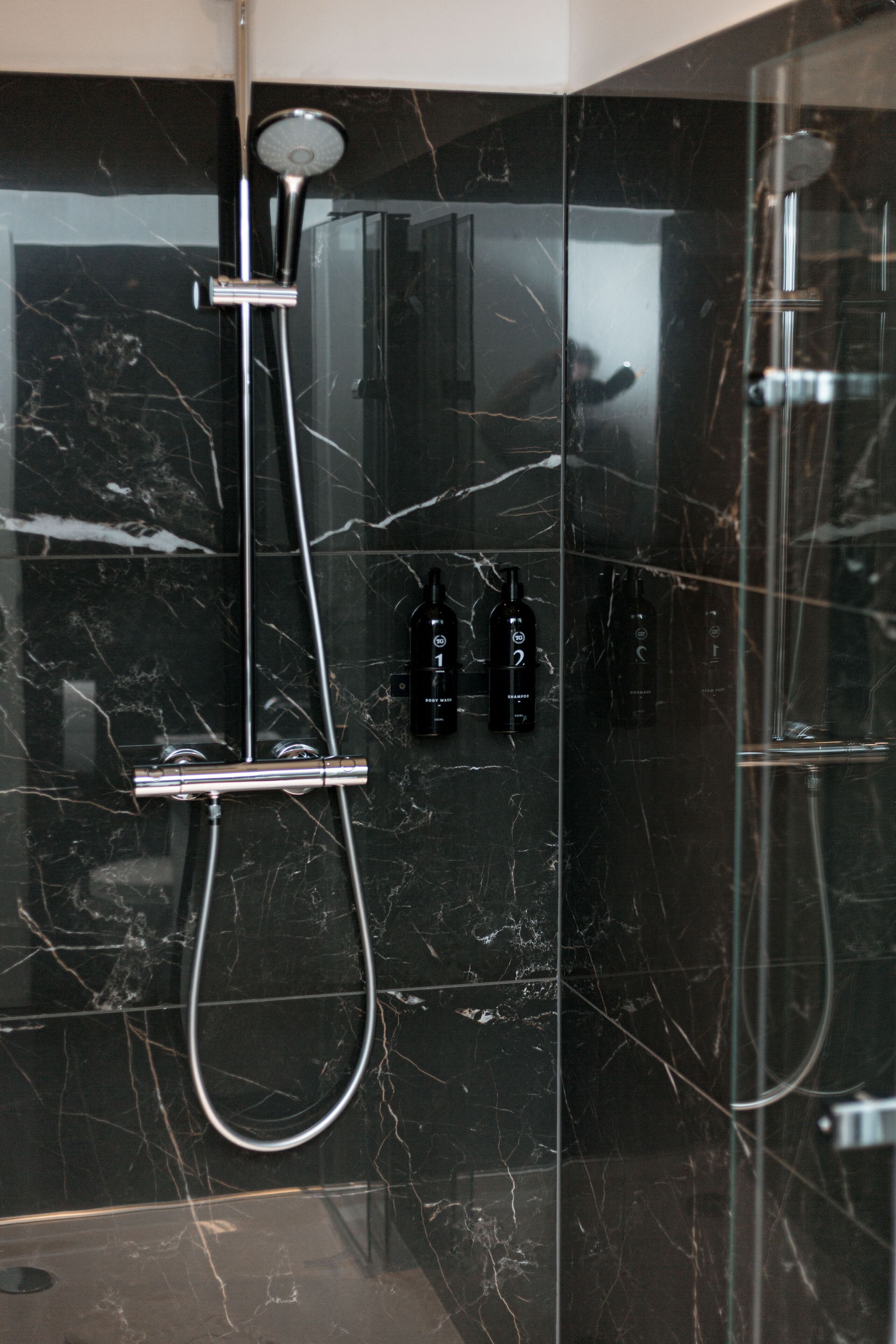 A modern bathroom with a black marble wall, a glass shower enclosure, and two bottles of shampoo.
