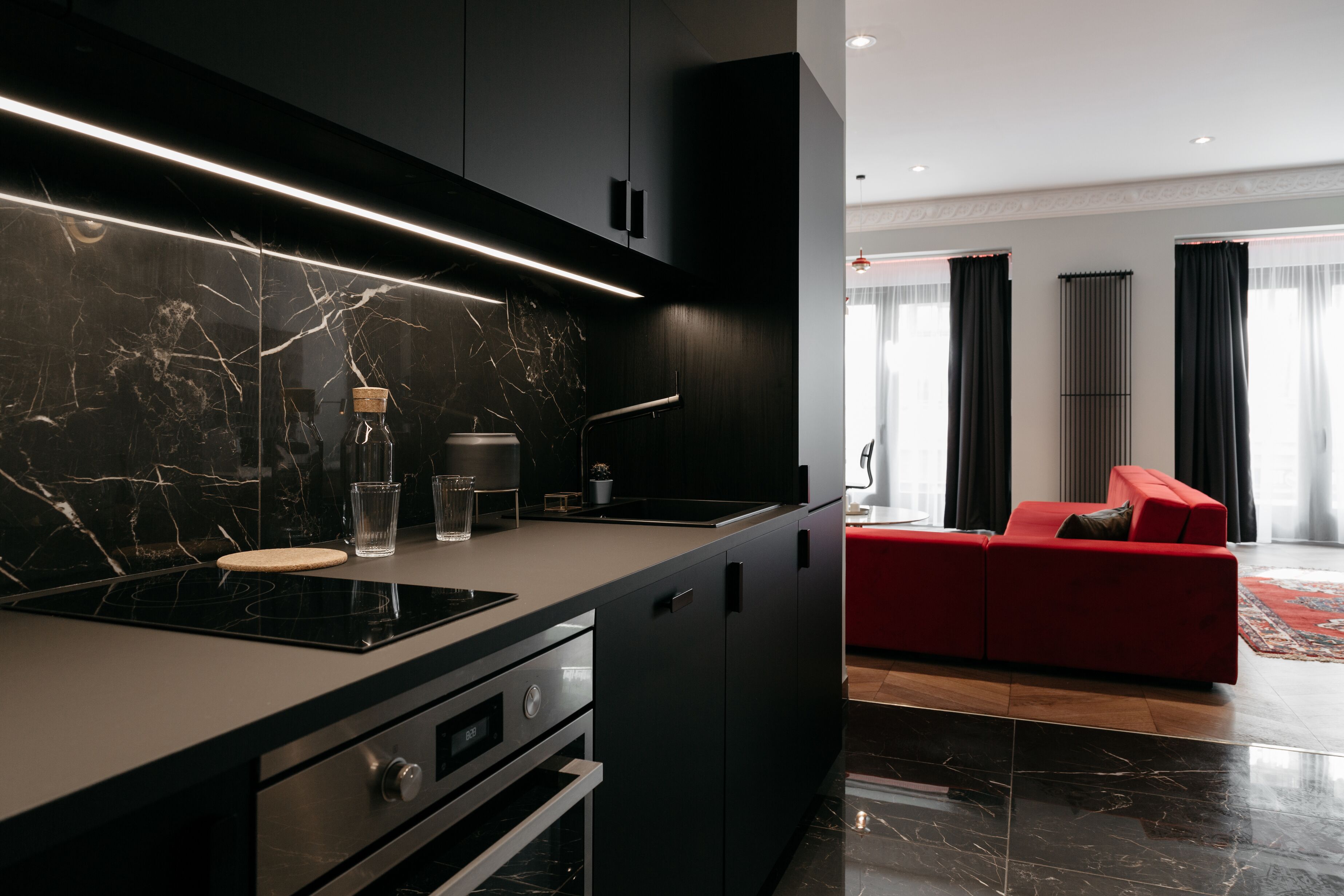 Une cuisine moderne avec un plan de travail noir, une cuisinière encastrée et un canapé rouge dans le salon attenant.