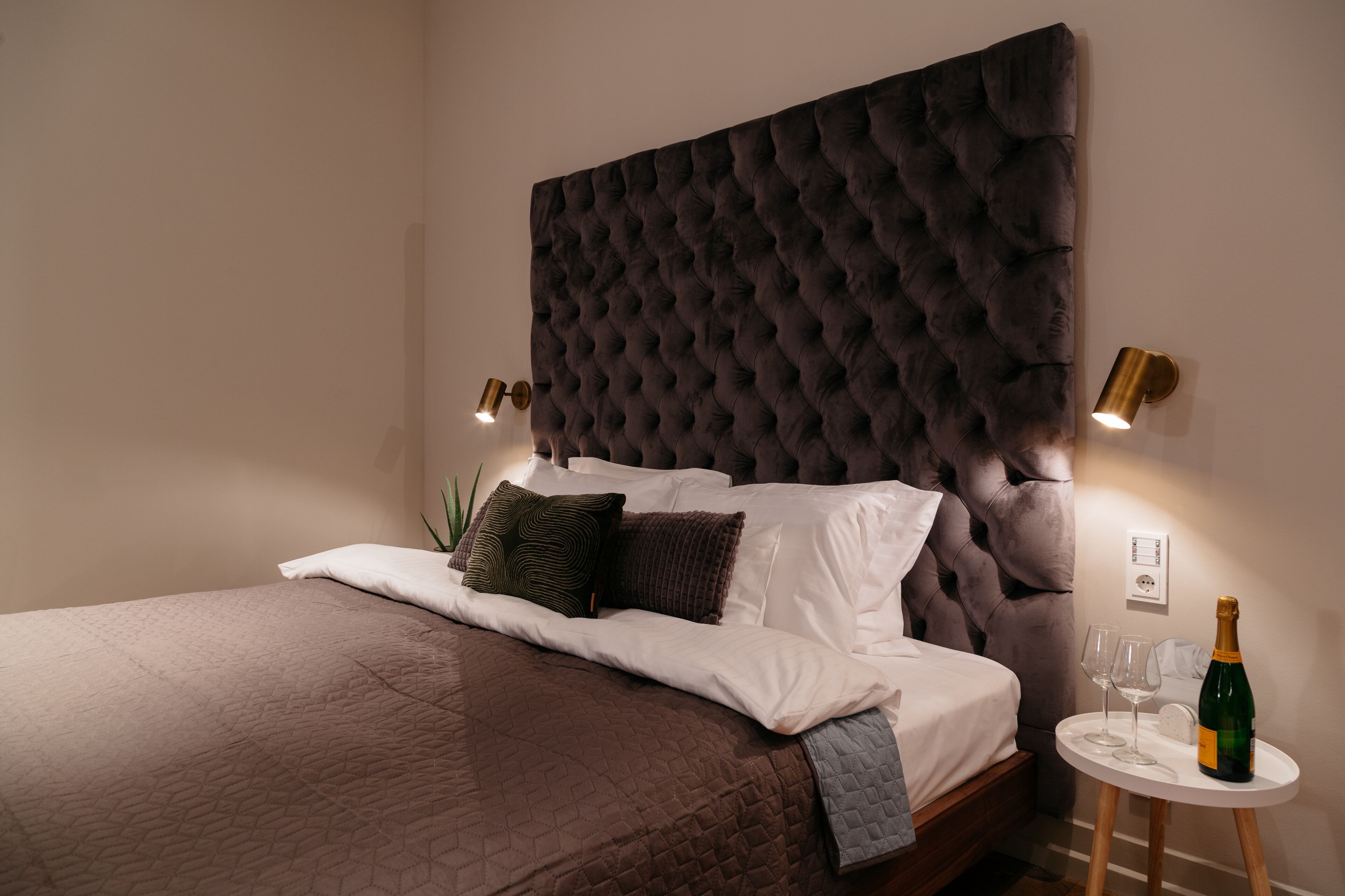 A neatly made bed with a tufted headboard, two bedside lamps, a small round table with a bottle and glasses, and a potted plant.