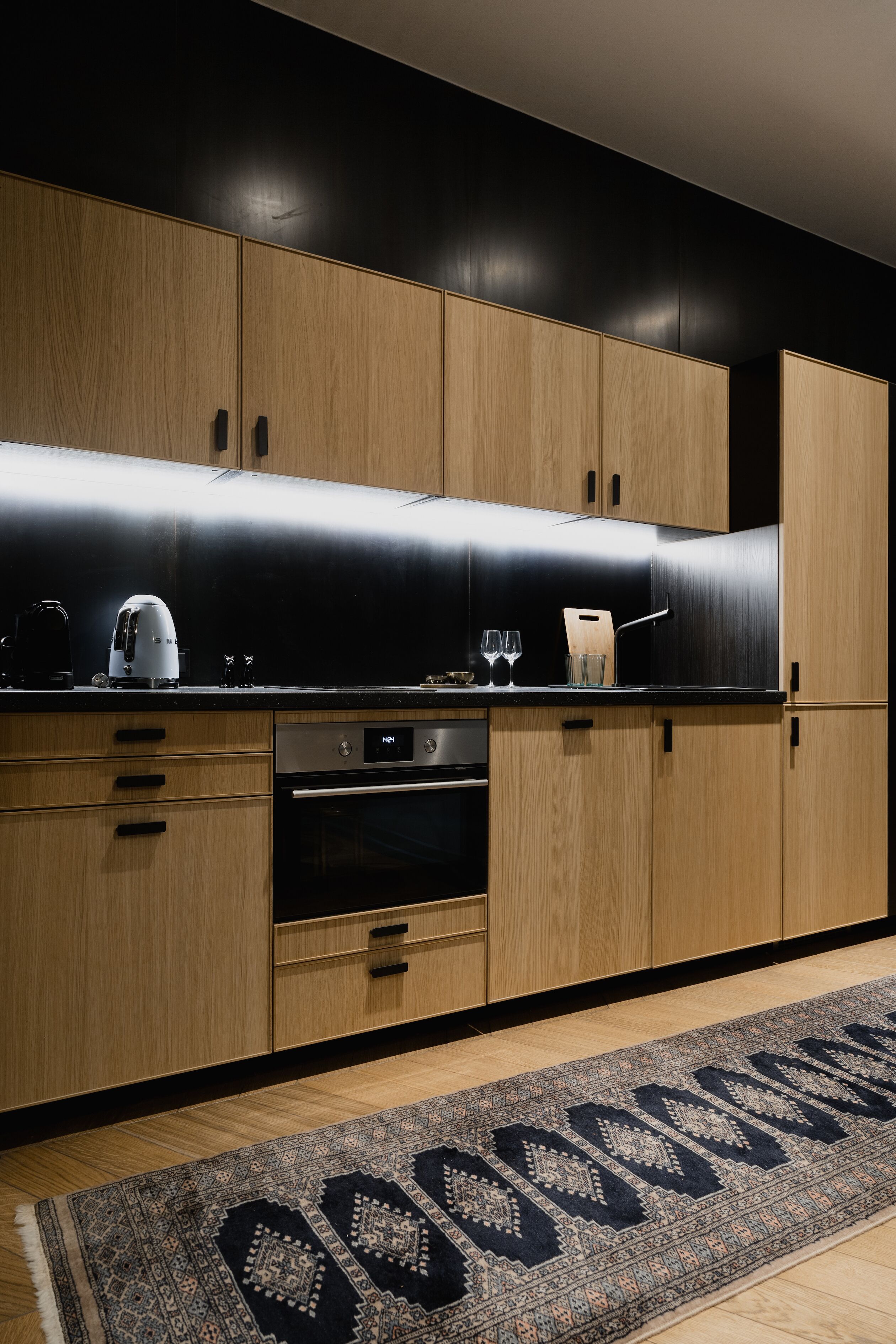 A modern kitchen with wooden cabinets, a black countertop, and a patterned rug.