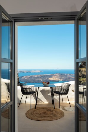 An open glass door leading to a terrace with a table and chairs, overlooking a coastal landscape. - Imera Vista Suites (Santorini)