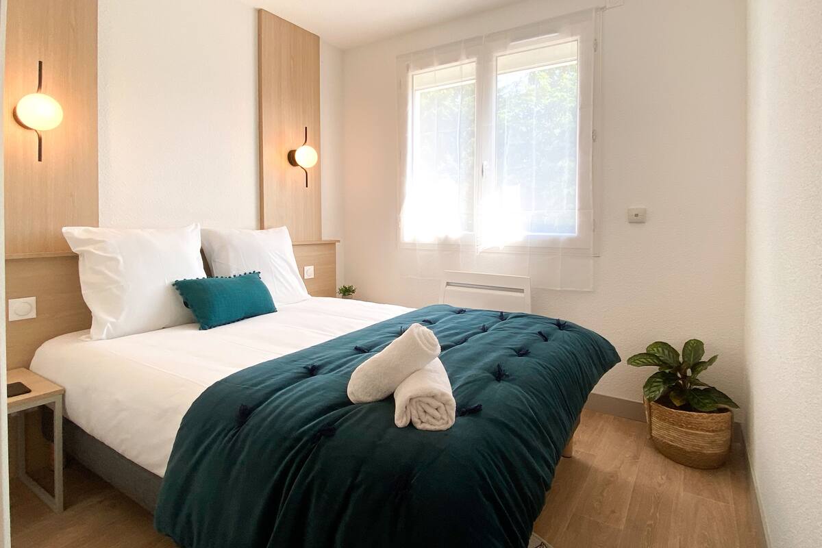 En pænt redt seng med en turkis sengetæppe og hvide puder, et natbord med en lampe, en potteplante og et vindue med gardiner.