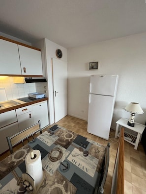 Cocina compacta con armarios blancos, horno empotrado y refrigerador. Hay una pequeña mesa con mantel corrido de temática náutica y una lámpara sobre una mesita.