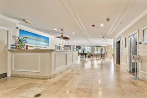 Un espace d’accueil d’h ôtel avec un comptoir sur lequel est affiché un panneau indiquant le « Waikiki Grand Hotel », un ventilateur de plafond et un panneau indiquant le deuxième étage.