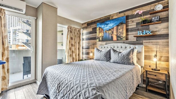 Een slaapkamer met een groot bed, een stadsgezichtschilderij aan de muur, een balkon met uitzicht op gebouwen en een houten plankje met de tekst 'Relax'.