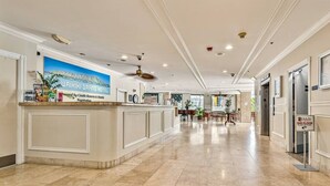 Un espace d’accueil d’hôtel avec un comptoir sur lequel est affiché un panneau indiquant le « Waikiki Grand Hotel », un ventilateur de plafond et un panneau indiquant le deuxième étage.