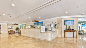 Een hotel receptie met een marmeren vloer, een receptiebalie, een zithoek met stoelen, een aan de muur bevestigde televisie en een lijst met een berglandschap.
