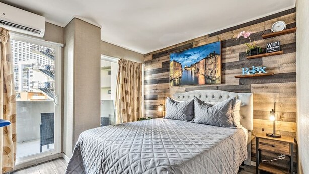 Une chambre avec un grand lit, un tableau représentant un paysage urbain accroché au mur, un balcon donnant sur les bâtiments et une étagère en bois sur laquelle est posée une pancarte indiquant « Détendez-vous ».