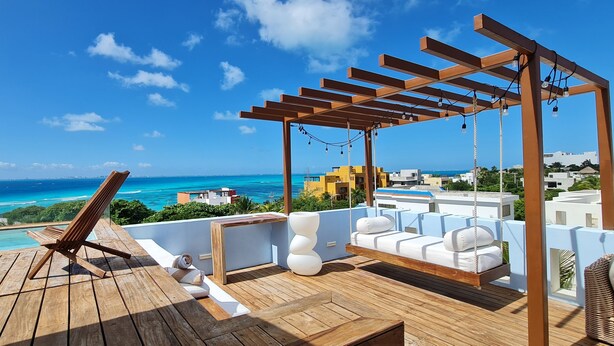 A wooden deck with a hammock, a swing, and a seating area with cushions, overlooking a clear blue sea and a coastal cityscape. - Casa Reyes at La Diosa - Stunning Roof Top Ocean view (Isla Mujeres)
