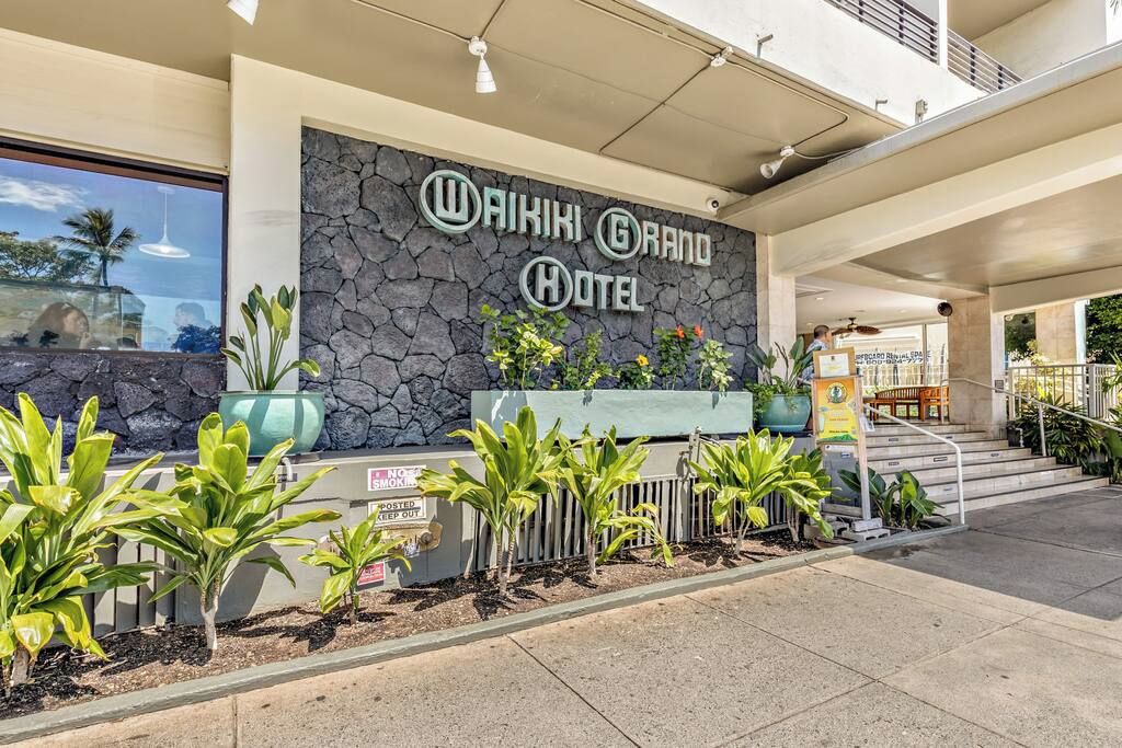 Het uiterlijk van het Waikiki Grand Hotel met een stenen gevel, een receptie en een trap naar de ingang.
