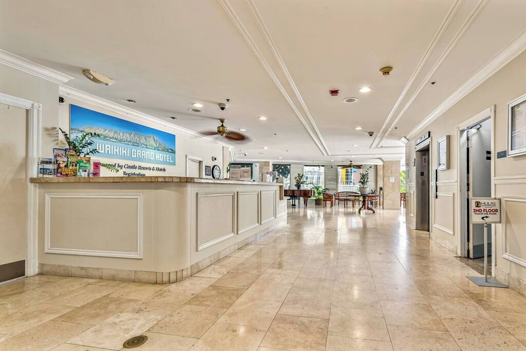 Un'area reception di un hotel con un bancone che mostra un cartello per il Waikiki Grand Hotel, una ventola a soffitto e una segnaletica che indica il secondo piano.