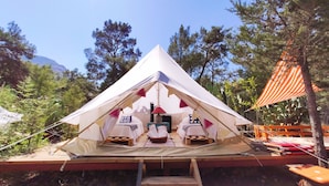 Una tienda de campaña triangular con dos camas y una mesa en el centro, rodeada de árboles y una montaña al fondo.
