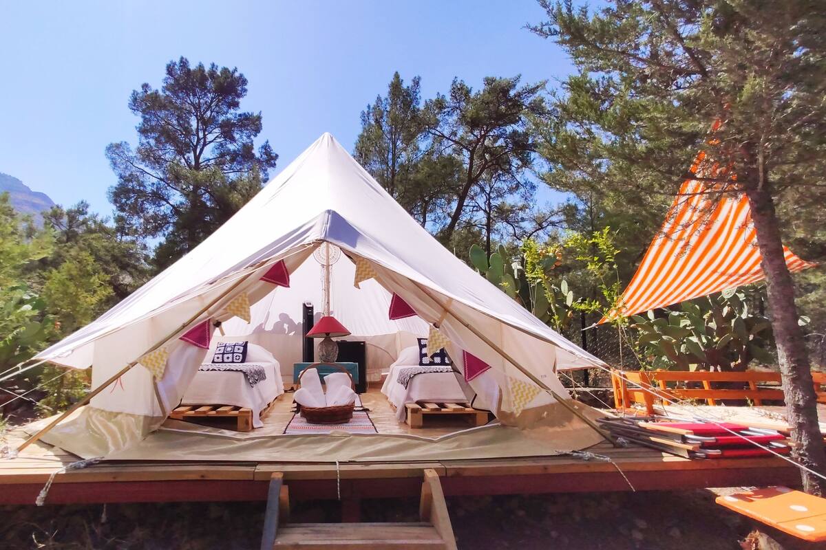 A triangular tent with two beds and a table in the center, surrounded by trees and a mountain in the background.