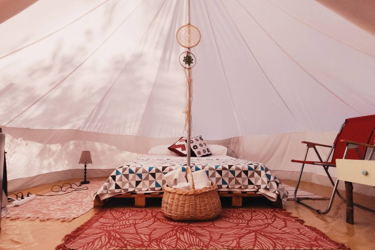 A tent with a bed, a red patterned rug, a wicker basket, a lamp, and a red chair.