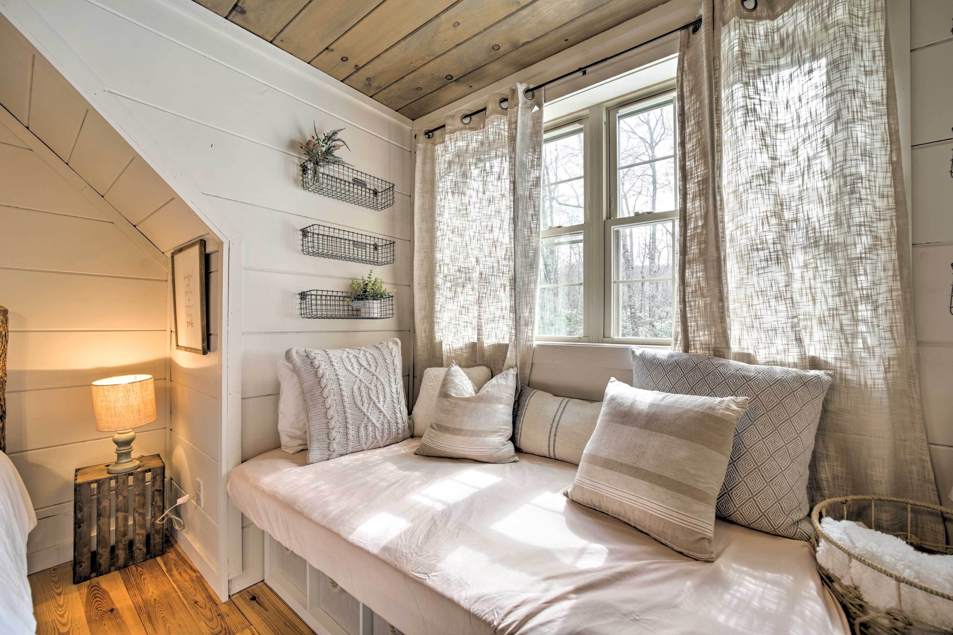 A cozy bedroom with a wooden ceiling, a bed with multiple pillows, a bedside lamp, and a window with sheer curtains.