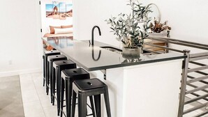 A modern kitchen with a central island, black bar stools, a potted plant, and a view of a desert landscape through a doorway. - Slick Rock Retreat House (Moab)