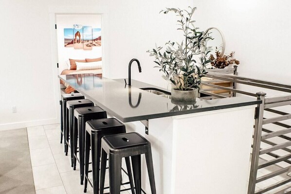 A modern kitchen with a central island, black bar stools, a potted plant, and a view of a desert landscape through a doorway. - Slick Rock Retreat House (Moab)